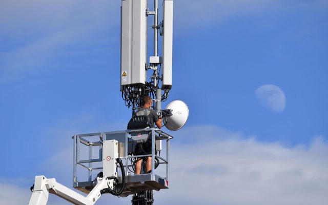 Communications technician in a raised bucket lift servicing a microwave dish and cellular antenna panels on a tower against a blue sky with the moon visible