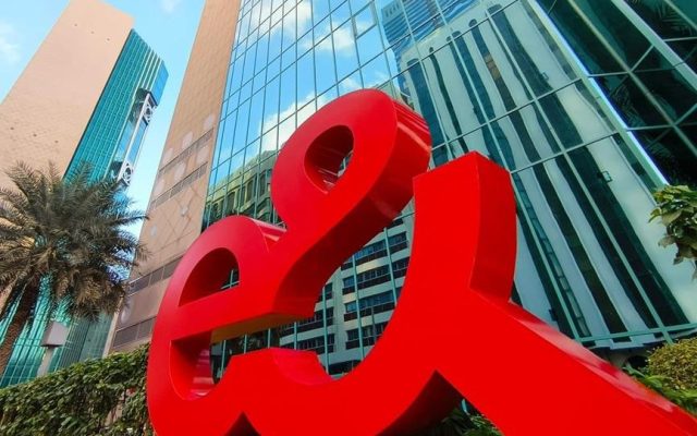 Large red three-dimensional e& logo sign outdoors in front of a modern glass tower with palm trees