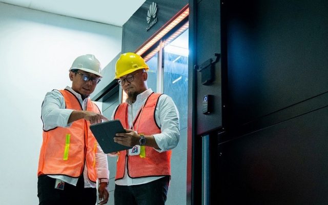 Two engineers in orange high-visibility vests and hard hats beside a Huawei equipment cabinet