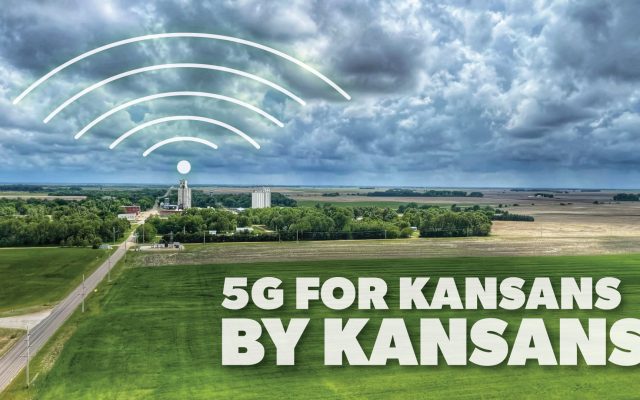 Aerial view of Kansas farmland with grain silos and a road under a dark cloudy sky, overlaid with a white signal icon and the words 5G FOR KANSANS BY KANSANS