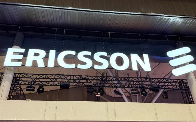 Illuminated Ericsson sign with the company logo hanging above a metal truss