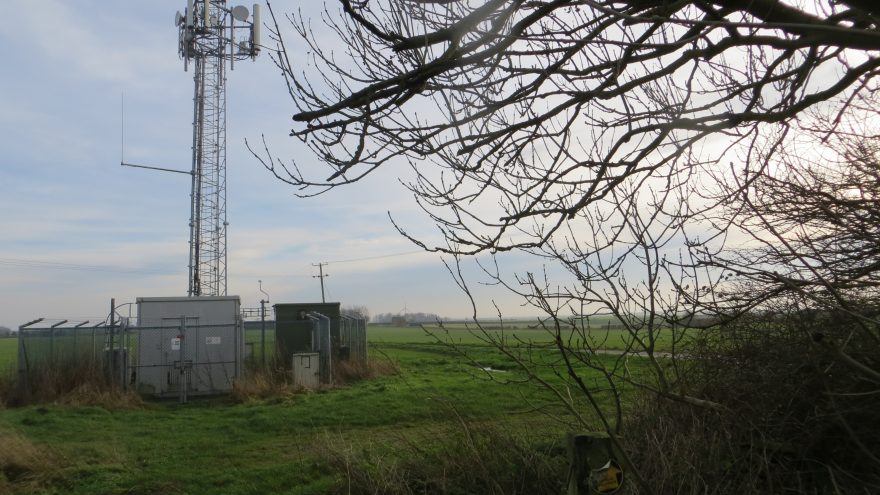 A lattice telecommunications mast with fenced equipment cabinets in a rural field